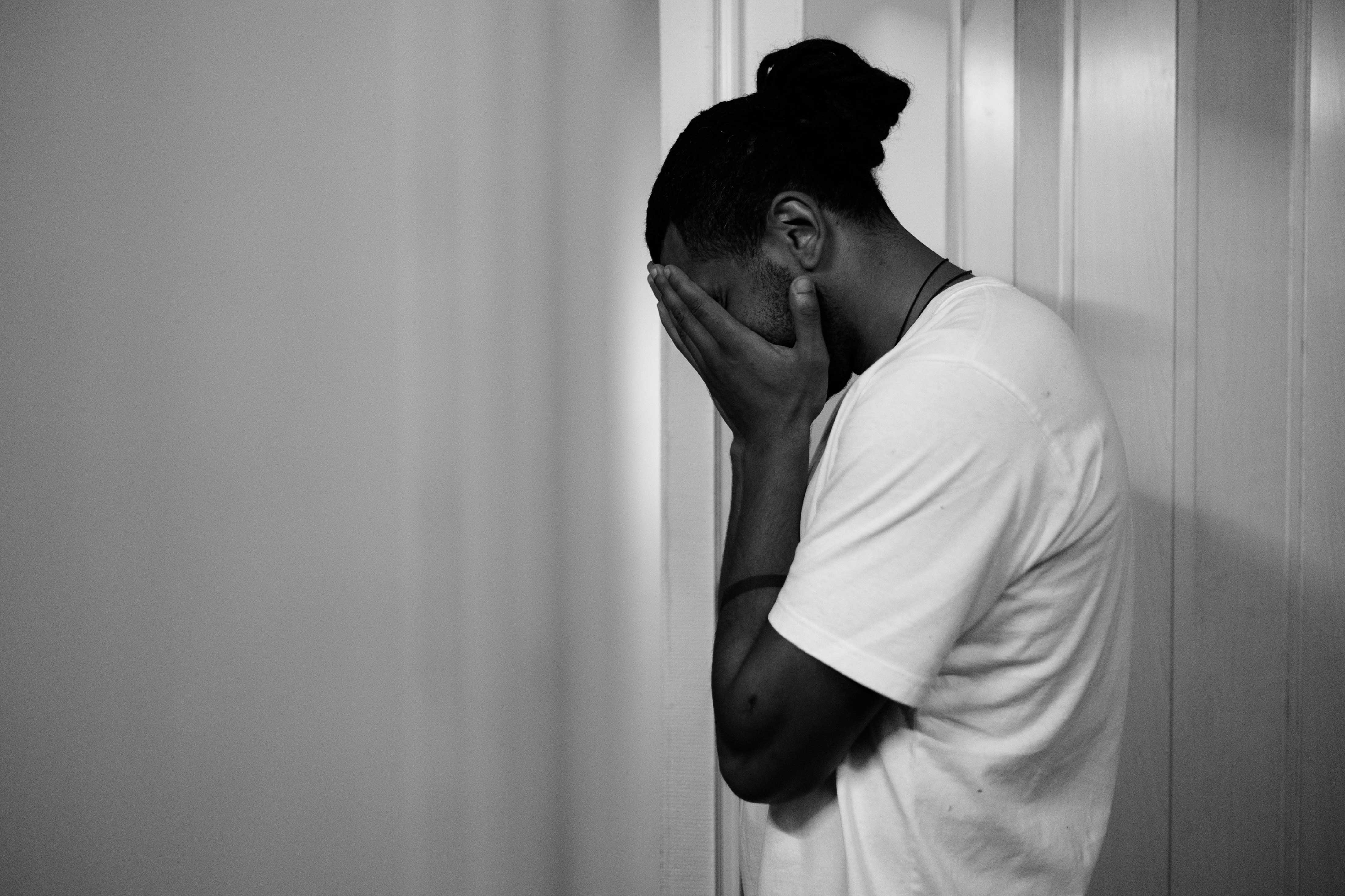 A person standing indoors, leaning against a wall with their face buried in their hands, appearing distressed or overwhelmed